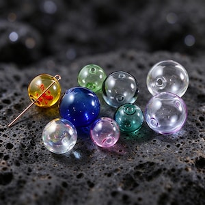 Könnte beinhalten: Eine Sammlung kugelförmiger Glasperlen in verschiedenen Farben, darunter transparent, blau, grün und gelb. Eine gelbe Perle enthält ein kleines rotes florales Element und wird von einer dünnen Metallnadel durchstochen.
