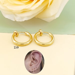以下が含まれることがあります： ゴールドトーンのクリップ式フープイヤリング2つ。イヤリングの直径は5.1cmです。イヤリングは、黄色いバラを背景にした白い表面に置かれています。イヤリングは、人の耳にも付けられています。
