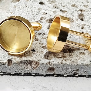 May include: A pair of gold cufflinks with round, smooth, blank tops. The cufflinks are made of metal and have a polished finish.