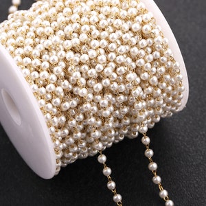 May include: A spool of gold chain with white pearl beads. The chain is wound around a white spool.