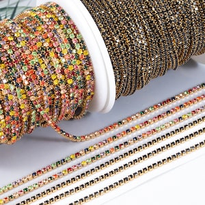 Puede incluir: Primer plano de dos carretes de cadena de strass. Un carrete presenta un arcoíris de colores, mientras que el otro carrete es negro. La cadena está hecha de pequeños strass cuadrados.