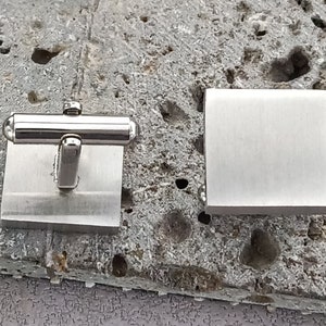 Puede incluir: Un par de gemelos de plata, uno con forma cuadrada y el otro con forma redondeada. Los gemelos están hechos de metal cepillado.