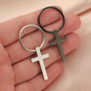 May include: Two silver and black cross keychains with a circular ring. The keychains are held in a hand.