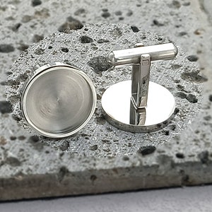May include: A pair of silver cufflinks with a round, smooth, silver surface. The cufflinks have a T-shaped bar that connects the two round surfaces.