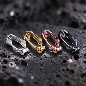 Könnte beinhalten: Vier metallene Clip-Ohrring-Konverter in Silber, Gold, Roségold und Schwarz. Jedes Teil verfügt über einen klappbaren Clip-Mechanismus mit einer kleinen Öse an der Basis zum Befestigen von Anhängern, konzipiert für nicht durchstochene Ohren.