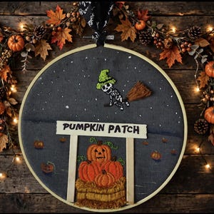 May include: A circular embroidery hoop featuring a dark gray fabric background with a stitched skeleton flying on a broomstick wearing a green hat. Below, a white sign reads PUMPKIN PATCH above a stack of embroidered orange pumpkins on hay.