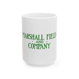 Op de afbeelding: Witte keramische mok met de groene tekst "MARSHALL FIELD AND COMPANY". De mok heeft een cilindrische vorm en een eenvoudig ontwerp, geschikt voor warme of koude dranken.