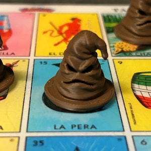 May include: A brown, 3D-printed, cone-shaped hat figurine with a wide brim and a folded, wrinkled texture resembling a face with deep-set eyes and a mouth. The object is positioned on a game board featuring colorful squares with text and illustrations.