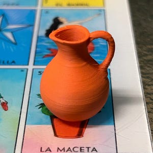 May include: A small, bright orange 3D-printed pitcher with a rounded body, a narrow neck, and a single curved handle. The surface features visible horizontal layer lines from the printing process. The pitcher is positioned on a game board.
