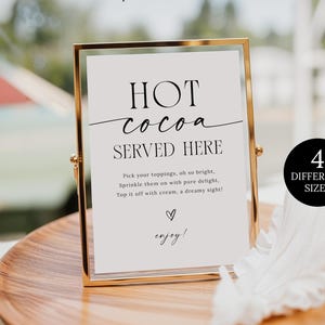 Peut inclure: Un panneau doré avec le texte "HOT cocoa SERVED HERE" sur fond blanc. Le panneau est posé sur un plateau en bois avec une plume blanche et un verre de liquide. Le panneau indique également "4 DIFFERENT SIZES".