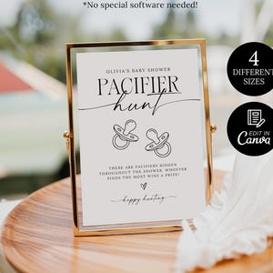 May include: A gold-framed sign for Olivia's baby shower, featuring the text "Pacifier Hunt" with pacifier illustrations. The sign is on a wooden tray with a white feather and a glass of liquid in the background. The sign is available in 4 different sizes and can be edited in Canva.