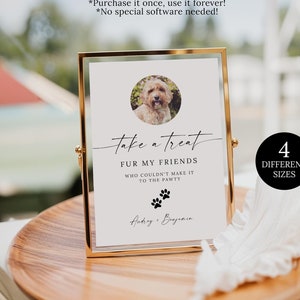 May include: A gold framed sign with a white background and black text that reads 'take a treat fur my friends who couldn't make it to the pawty' with a paw print graphic. The sign also has a photo of a dog in a circle. The text '4 different sizes' is in a black circle on a white background. The sign is on a wooden table with a white feather and a glass of brown liquid.