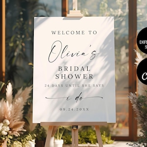 May include: A white sign on a wooden easel with the text "Welcome to Olivia's Bridal Shower." The sign also includes the text "24 days until she says I do" and the date "09.24.20XX."