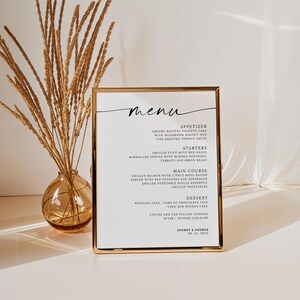 Op de afbeelding: Een goudomrand menu met sierlijk handschrift, met gerechten zoals voorgerechten en desserts. Links staat een vaas met gedroogde tarwestengels. Het menu staat op een wit oppervlak.