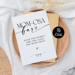 May include: Printable sign for a "Mom-osa Bar" with instructions to "Pour the bubbly, add the juice, top with fruit" and the word "enjoy" in cursive. The sign is on a wooden surface with a brown envelope and a white background.