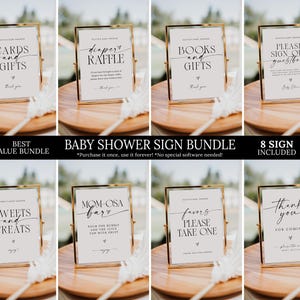 May include: A collection of eight baby shower signs in gold frames, displayed on a wooden tray. Each sign features elegant calligraphy with phrases like "Cards and Gifts" and "Diaper Raffle." The signs are part of a "Baby Shower Sign Bundle."