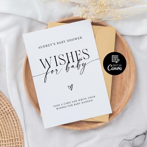 以下が含まれることがあります： 白地に黒文字で「Audrey's Baby Shower Wishes for Baby」と書かれたカード。ハートマークと「Take a card and write your wishes for Baby Hanson.」というテキストも書かれている。カードは木製の表面に置かれ、茶色の封筒と、白文字で「Edit in Canva」と書かれた黒い円形がある。