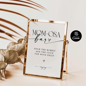 May include: A gold-framed sign with the text "MOM-OSA bar" in black cursive font. The sign also includes the instructions "POUR THE BUBBLY ADD THE JUICE TOP WITH FRUIT" and the word "enjoy" in a smaller font.