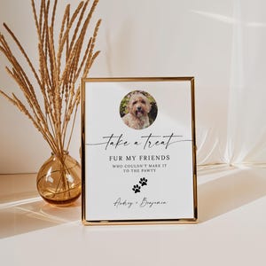 May include: A white sign with black text that reads "Take a treat fur my friends who couldn't make it to the pawty."  The sign has a photo of a dog in the top left corner and paw prints below the text. The sign is in a gold frame.