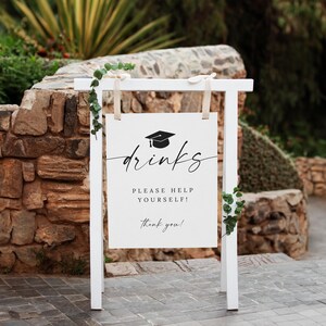 May include: A white wooden sign with the word "drinks" in elegant script, topped with a graduation cap icon. The sign reads "Please help yourself! Thank you!" and is decorated with greenery.