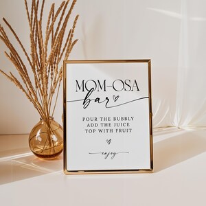 May include: A gold-framed sign with the text "MOM-OSA bar" in black cursive font. The sign also includes instructions for making a mimosa: "POUR THE BUBBLY ADD THE JUICE TOP WITH FRUIT" and the word "enjoy" in cursive.
