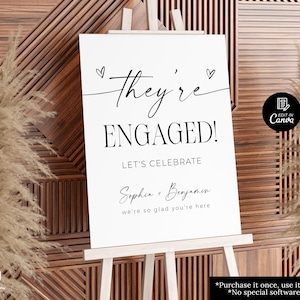 Puede incluir: Un letrero blanco sobre un caballete de madera con las palabras "They're ENGAGED!" en escritura negra. Debajo, se lee "LET'S CELEBRATE" y los nombres "Sophia + Benjamin". El letrero está decorado con pequeñas ilustraciones de corazones.