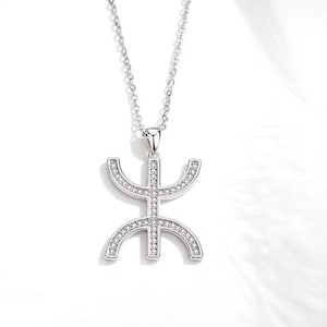 May include: A silver necklace with a pendant in the shape of a stylized symbol. The pendant is covered in small, clear cubic zirconia stones.