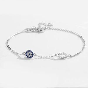 Puede incluir: Un brazalete de cadena de plata con un dije de ojo azul malvado y un dije de mano de hamsa. Ambos dijes están decorados con piedras de circonita cúbica transparentes.