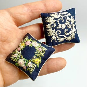 Puede incluir: Dos cojines cuadrados en miniatura de color azul marino con diseños florales bordados en blanco. Uno presenta una corona de flores rosas, amarillas y blancas. El otro tiene un diseño floral blanco en espiral. Ambos cojines tienen ribete blanco.