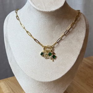 May include: Gold chain necklace with a green clover charm, a green square charm, and a gold charm with a letter B and clear rhinestones.