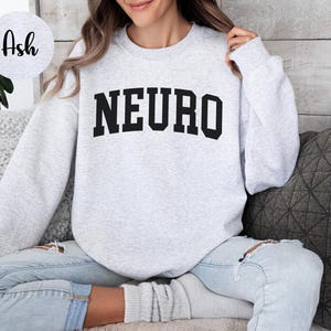 K&ouml;nnte beinhalten: Hellgraues Sweatshirt mit dem Wort "NEURO" in fetten, schwarzen Buchstaben auf der Brust. Das Sweatshirt hat einen Rundhalsausschnitt und lange &Auml;rmel. Das Model tr&auml;gt eine hellblaue Jeans.