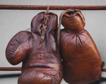 Puede incluir: Un par de guantes de boxeo vintage de cuero marrón colgados de una barra de madera. Los guantes muestran signos de desgaste, con cierres con cordones y una apariencia desgastada, lo que sugiere su uso en boxeo o entrenamiento.