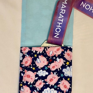 May include: A blue fabric pouch with a navy blue flap featuring a pattern of pink pigs with wings, clouds, and stars. A marathon medal is visible, with a purple ribbon that reads "MARATHON".
