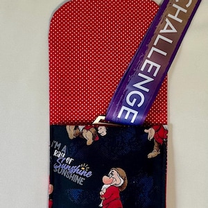 May include: A fabric pouch featuring a red and white polka dot flap and a black main body printed with the character Grumpy from Snow White, the text "I'M A RAY OF SUNSHINE", and a purple lanyard with the visible text "EY CHALLENGE" attached.