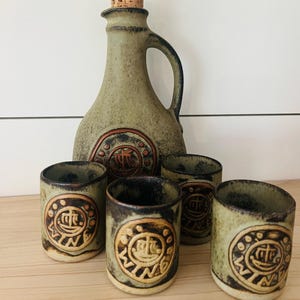 May include: A vintage ceramic liquor set featuring a green, textured decanter with a cork stopper and a handle, accompanied by five matching shot glasses. Each piece is adorned with a circular, embossed design in a contrasting color.