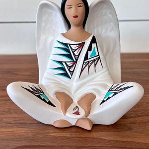 May include: A white ceramic angel figurine with black hair, closed eyes, and red lips. The angel is seated with legs crossed, wearing a white robe with black, turquoise, and red geometric designs. White wings extend from the back.