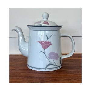 May include: White ceramic teapot with a lid, featuring a floral design in pink and purple. The teapot has a curved spout and a handle. The floral pattern is complemented by gray accents. A classic piece for serving tea.