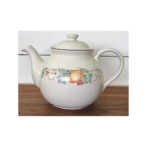 May include: Cream-colored ceramic teapot with a lid and handle. The teapot is decorated with a fruit and leaf design in shades of orange, green, and red. The rim of the teapot is trimmed with a thin brown line.