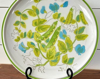 Może przedstawiać: Biały ceramiczny talerz z zieloną obwódką, z botanicznym wzorem zielonych strąków groszku, niebieskich kwiatów oraz zielonych i żółtych liści. Talerz jest prezentowany na czarnym metalowym stojaku.