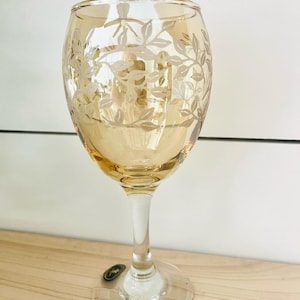 May include: A stemmed wine glass with a light amber bowl and clear stem and base. The bowl features a delicate etched floral design. The glass is set on a light-colored wooden surface.