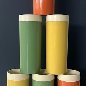 May include: A stack of six cylindrical tumblers in shades of orange, yellow, and green, each with a cream-colored rim. The tumblers are arranged in a pyramid shape against a dark blue background.