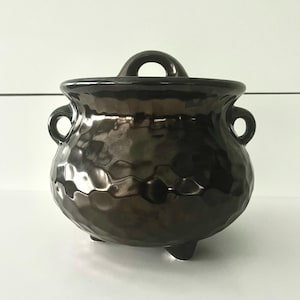 May include: A dark brown, ceramic cauldron with a textured surface. The cauldron has a rounded body, three small feet, and two loop handles on the sides. A larger loop handle is on top. The cauldron is likely used for decorative purposes.