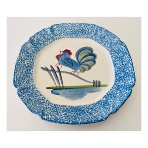 May include: A ceramic plate with a blue and white speckled rim. The center features a blue rooster perched on a branch above a blue pond. The rooster has a red comb and a green and blue tail.