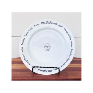 May include: White ceramic plate with an illustration of a person wearing glasses and a hat. The plate is encircled with the words "Old Fashioned", "sugar", "orange bitters", "water", "bourbon", "lemon twist", and "cherry".