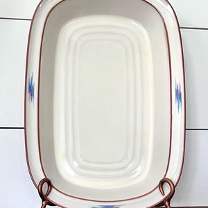 May include: A white ceramic baking dish with a red and blue geometric pattern around the rim. The dish is on a metal stand with two hooks.