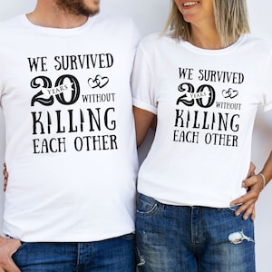 K&ouml;nnte beinhalten: Zwei wei&szlig;e T-Shirts mit schwarzem Text, der lautet "Wir haben 20 Jahre &uuml;berlebt, ohne uns gegenseitig umzubringen".