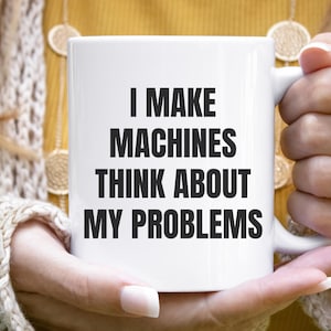 May include: A white ceramic mug featuring the black printed text I MAKE MACHINES THINK ABOUT MY PROBLEMS. The mug has a standard cylindrical shape with a side handle and a smooth finish, displaying the bold sans-serif lettering clearly on the front.