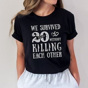 K&ouml;nnte beinhalten: Schwarzes T-Shirt mit wei&szlig;em Text, der "We survived 20 years without killing each other" lautet. Der Text ist mit einer Herzgrafik und einem Banner mit der Aufschrift "years" gestaltet.