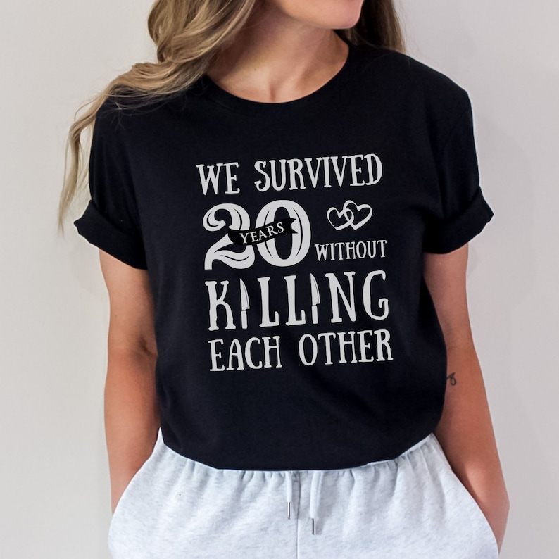 K&ouml;nnte beinhalten: Schwarzes T-Shirt mit wei&szlig;em Text, der "We survived 20 years without killing each other" lautet. Der Text ist mit einer Herzgrafik und einem Banner mit der Aufschrift "years" gestaltet.