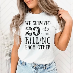 K&ouml;nnte beinhalten: Wei&szlig;es T-Shirt mit schwarzem Text, der "We survived 20 years without killing each other" lautet. Die Zahl 20 ist mit einem Banner mit der Aufschrift "years" gestaltet und zwei Herzen befinden sich &uuml;ber dem Wort "without".